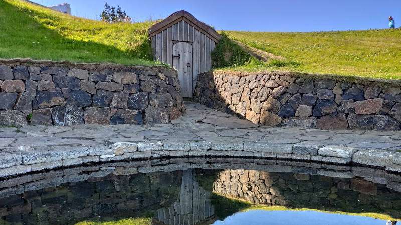 Reykholt icelandic style shed with stone walls and a small pond in the front on a sunny day