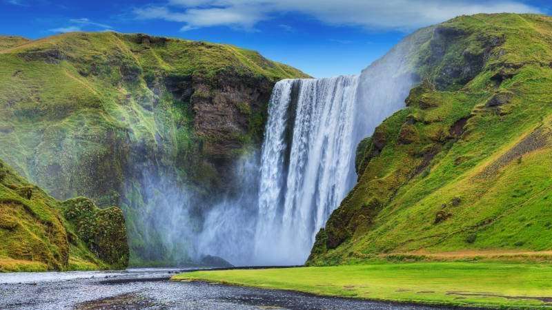 Skogafoss waterfall on a sunny day with green hills around